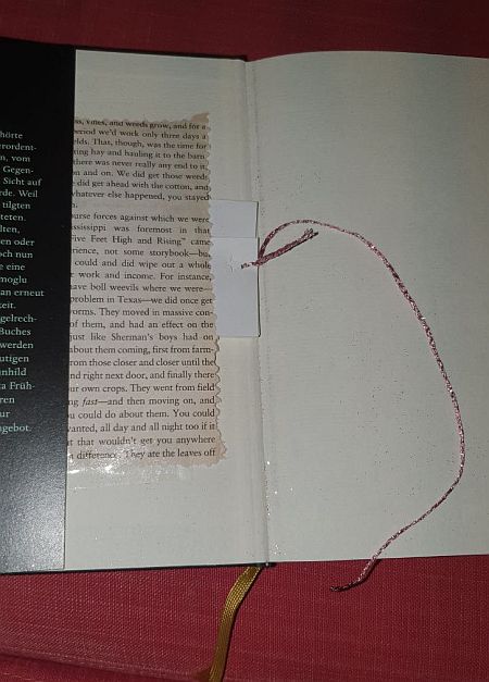 Aufgeklapptes Buch. Unter dem Schutzumschlag lugt ein festgeklebter Zeitungsausschnitt hervor, der eine kleine Tasche bildet. Aus der Tasche lugt ein Glitzfaden hervor. Das ist das Buchorakel aus meiner Buchhandlung in unserm Veedel