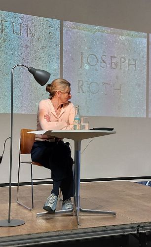 Frau in rosa Pull an Tisch mit Lampe neben sich, hinter ihr Fotos der Bodenplatten mit dem Namenszug Joseph Roth und den den letzten Buchstaben von "Keun".